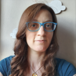 A photo of Bailey—she has long, curled hair; is wearing the brightest blue frames you can imagine and a teal shirt; and may-or-may-not be smiling (it's hard to tell). Behind her on the wall are some clouds (made of cork, painted white).